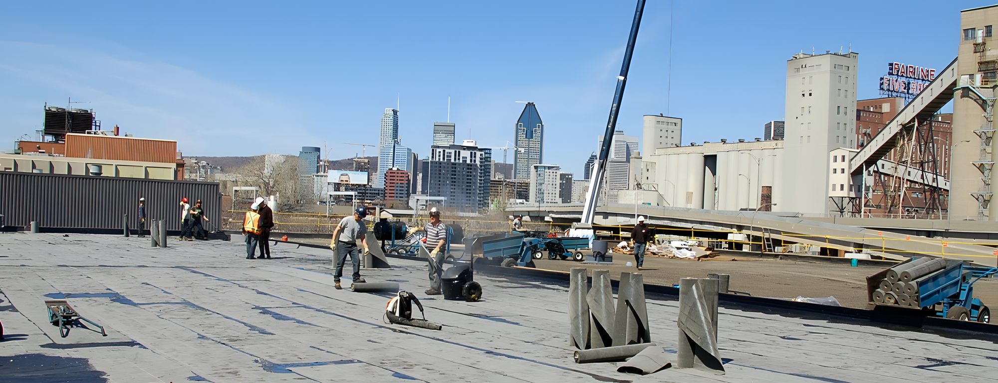 Un grand toit plat en chantier, une grue, l'équipe au travail, les tours du centre-ville de Montréal à l'horizon