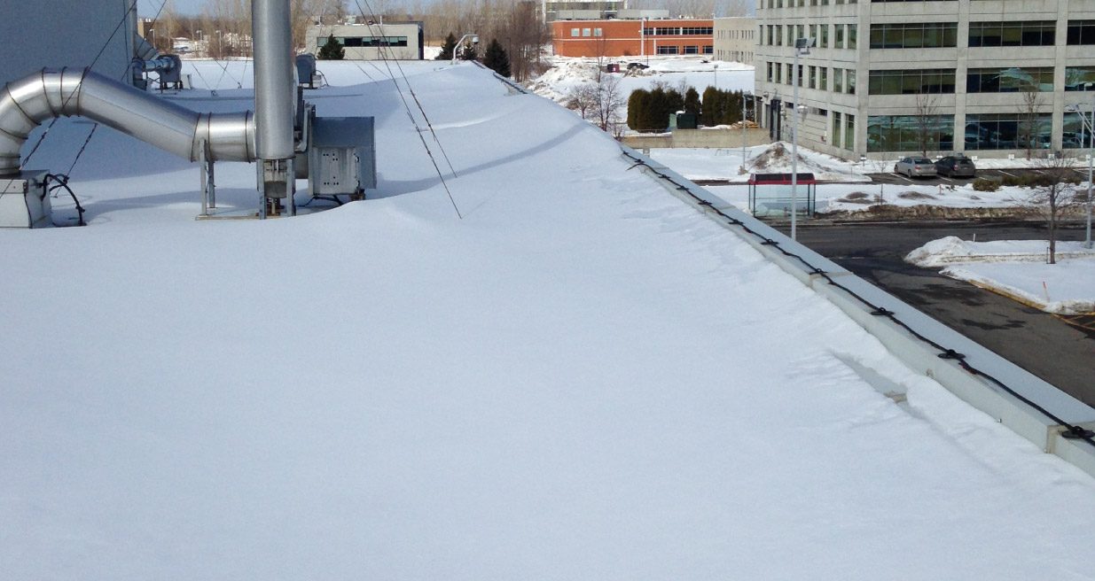 Un toit plat couvert de neige, un conduit dépasse, des tours de bureaux au loin