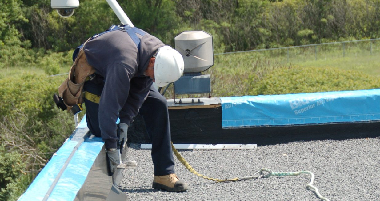 Un couvreur casqué, harnais au dos, inspecte le rebord d'un toit plat gravillonné